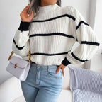 Woman wearing a white and black striped knit sweater with a high neckline and long sleeves, holding a white purse, paired with light blue jeans.