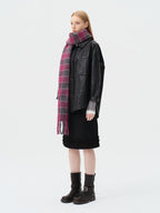 Side view of a woman wearing a black leather-look jacket, a black skirt with ruffled hem, a pink and grey plaid scarf, and black boots.