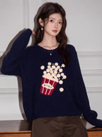 Woman wearing a navy blue knit sweater with a popcorn graphic and Korean text, touching her shoulder.