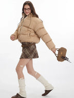 Model walking while wearing a tan cropped puffer jacket, brown plaid pleated mini skirt, white leg warmers, and brown loafers.