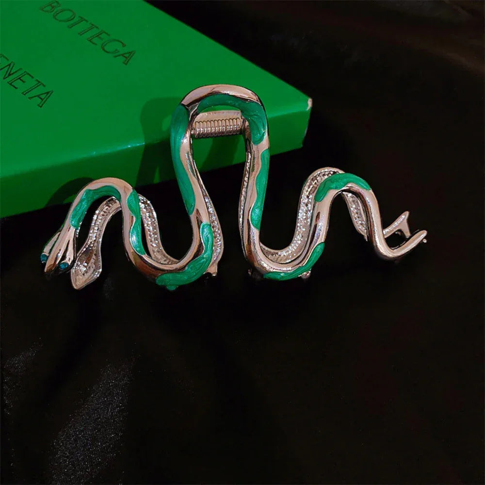 Snake-shaped hair clip with green and silver enamel on a black surface next to a green box.