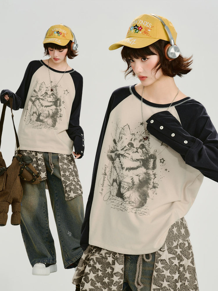 Full body shot of a model wearing a cream and navy raglan long-sleeve shirt with a cat graphic print, layered over a patterned skirt and jeans, with a yellow cap and headphones.
