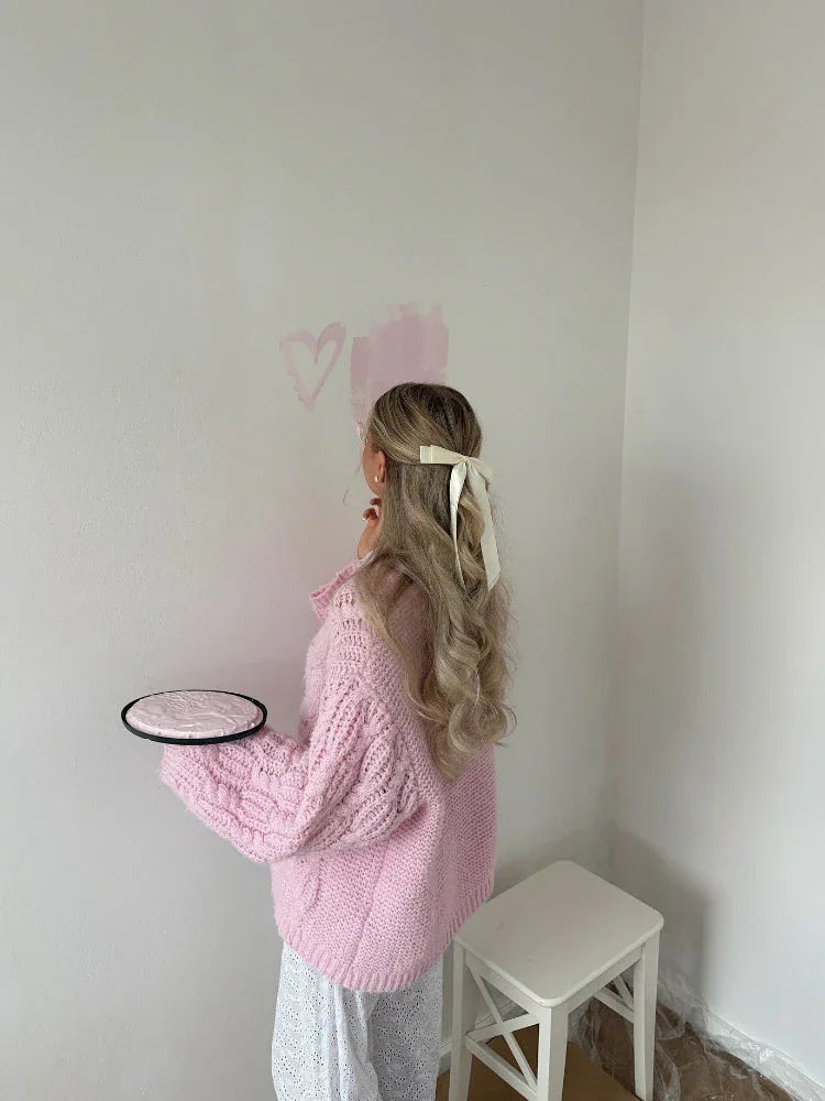Woman with blonde hair in a bow, wearing a pink cable knit sweater and white dress, holding a pink circular object, with hearts painted on a wall.