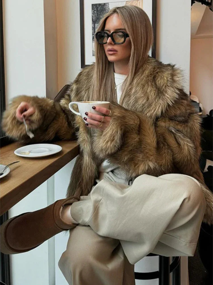 Woman in a brown faux fur jacket, beige pants, and sunglasses, holding a cup of coffee.