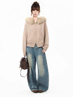 Woman wearing a beige knit cardigan with a faux fur collar and wide-leg blue jeans, holding a brown backpack