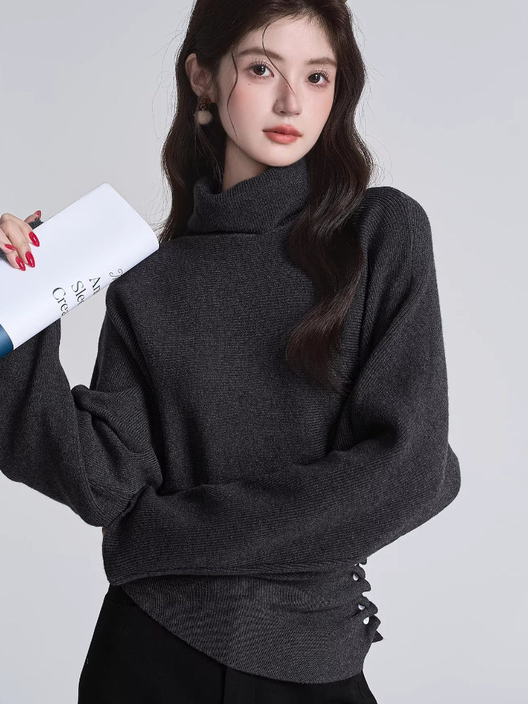 Woman wearing a dark grey turtleneck sweater with batwing sleeves and a cinched button detail at the waist, holding a book.