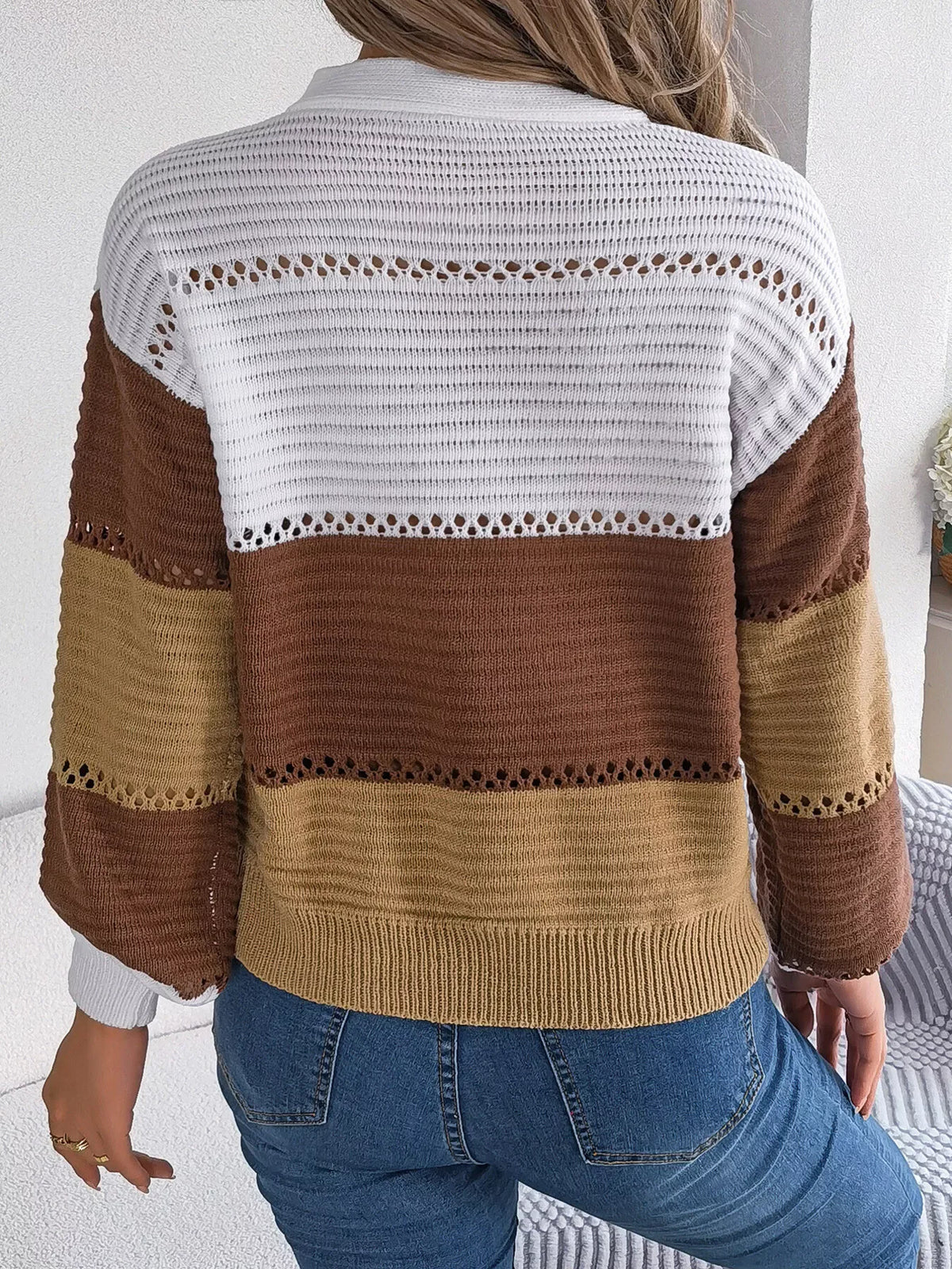Back view of a woman wearing a brown, tan, and white color block knit sweater with a v-neck and button details, paired with blue jeans.