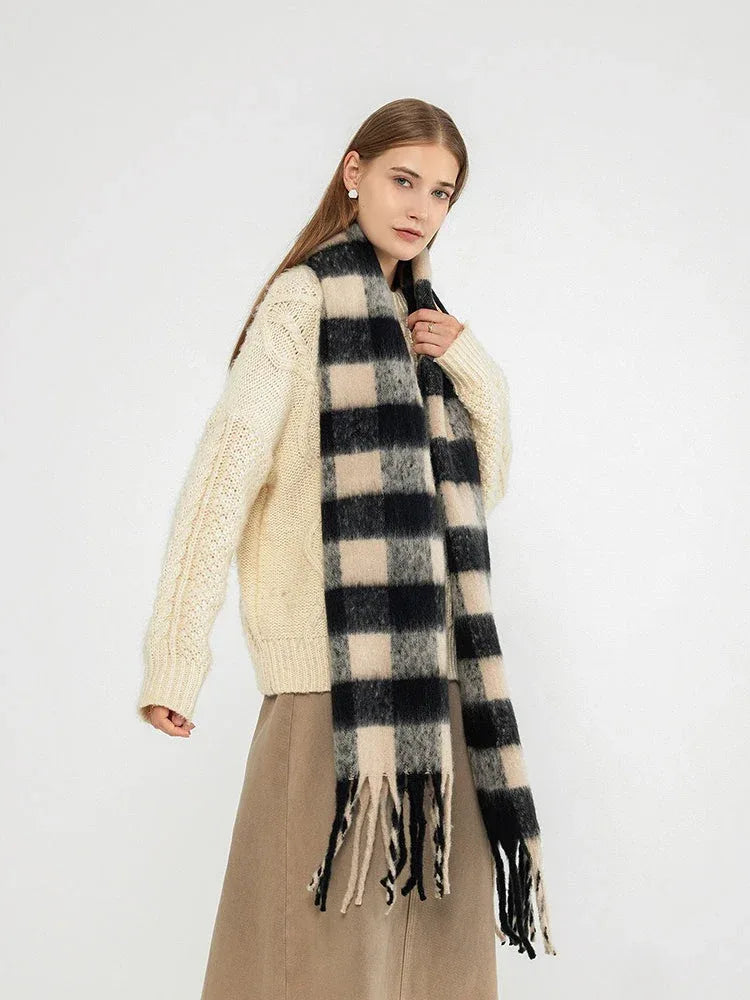 Woman wearing a cream cable knit sweater and a large black and beige plaid scarf with fringe, posing with one hand on her hip.