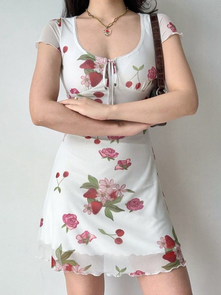 Woman wearing a white short-sleeve dress with a strawberry and rose print, featuring a tie-front neckline and a sheer hem.