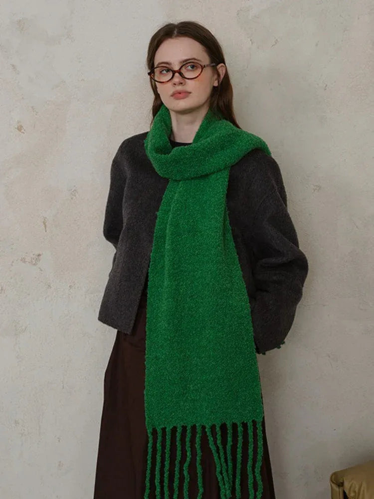 Woman wearing a dark grey sweater and a bright green textured knit scarf with fringe, standing against a textured wall.