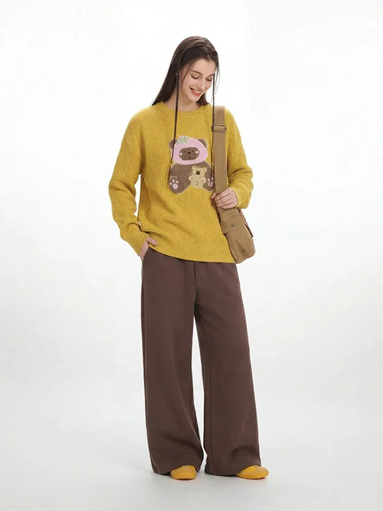 Woman smiling and wearing a yellow crew neck sweater with a bear graphic and brown wide-leg pants.