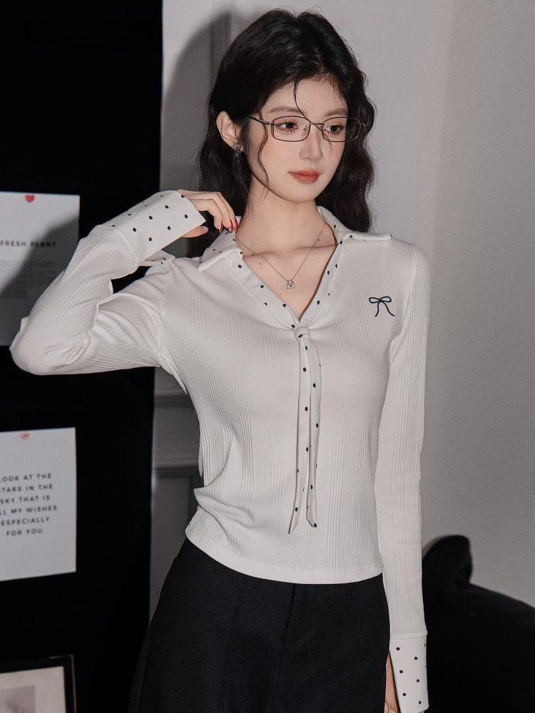 Woman posing with her hand touching her hair, wearing a white ribbed long-sleeve shirt with polka dot accents.