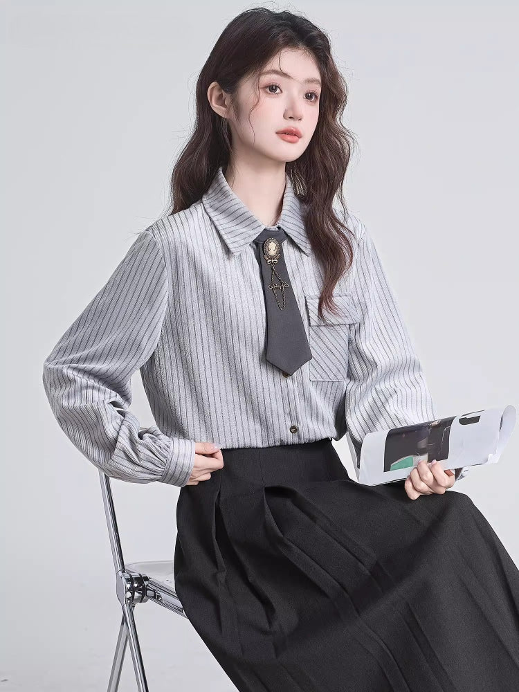 Woman sitting on a chair wearing a grey pinstripe collared shirt, black pleated skirt, and holding a folded magazine.
