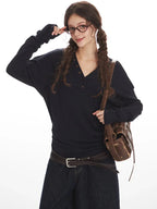 Close-up of a woman wearing a dark long-sleeved V-neck top, adjusting her glasses, with a brown leather crossbody bag, posing against a white background.