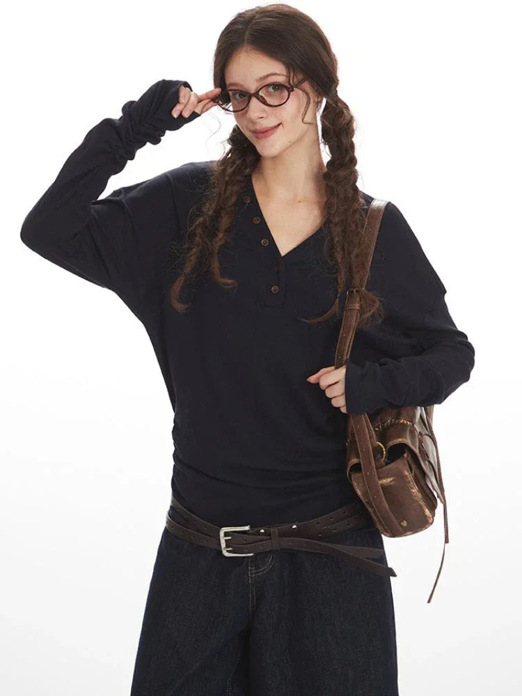 Close-up of a woman wearing a dark long-sleeved V-neck top, adjusting her glasses, with a brown leather crossbody bag, posing against a white background.