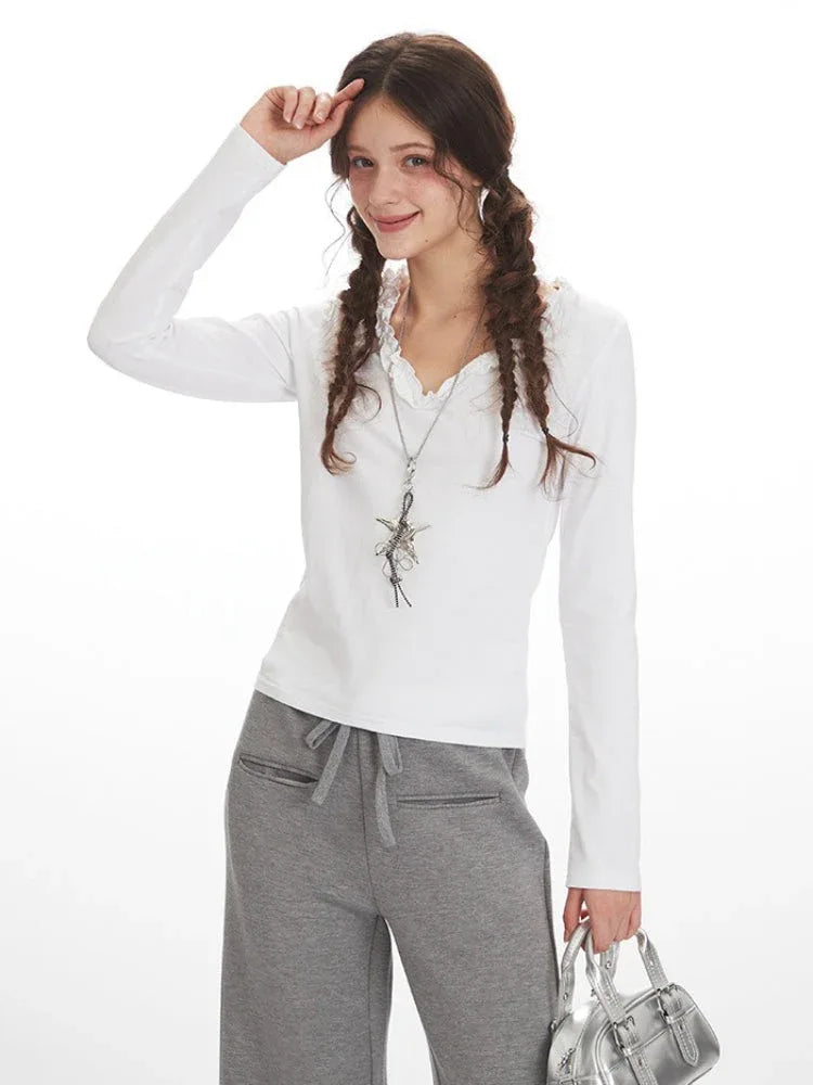 Close-up of a woman wearing a white long-sleeve shirt with ruffled neckline and grey sweatpants, posing with hand on her head.