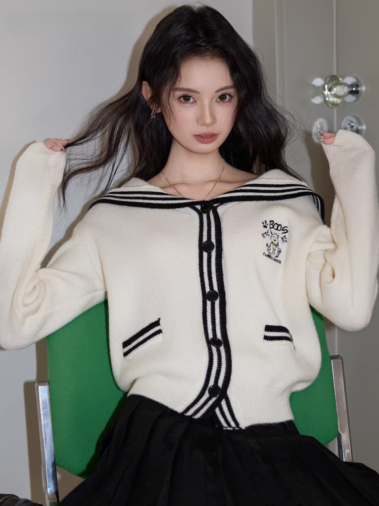Full body shot of a woman wearing a white knit cardigan, black skirt, and black shoes, standing next to a green chair.