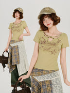 Full-body shot of a model wearing a light green short-sleeve t-shirt with a cutout neckline and star details, paired with an asymmetrical plaid skirt and a khaki baseball cap, holding a brown handbag. A smaller inset image shows a close-up of the outfit.