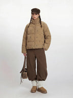 Woman wearing a tan corduroy puffer jacket with a repeating dog print, brown wide-leg pants, and a brown cap, holding a brown leather bag.