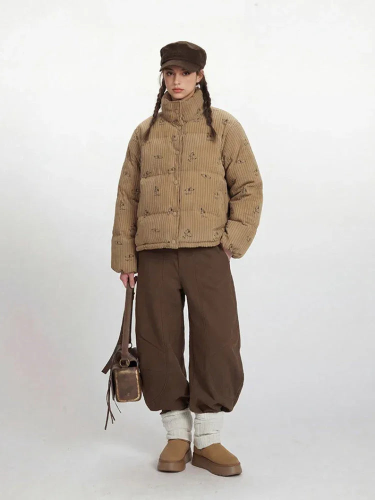 Woman wearing a tan corduroy puffer jacket with a repeating dog print, brown wide-leg pants, and a brown cap, holding a brown leather bag.