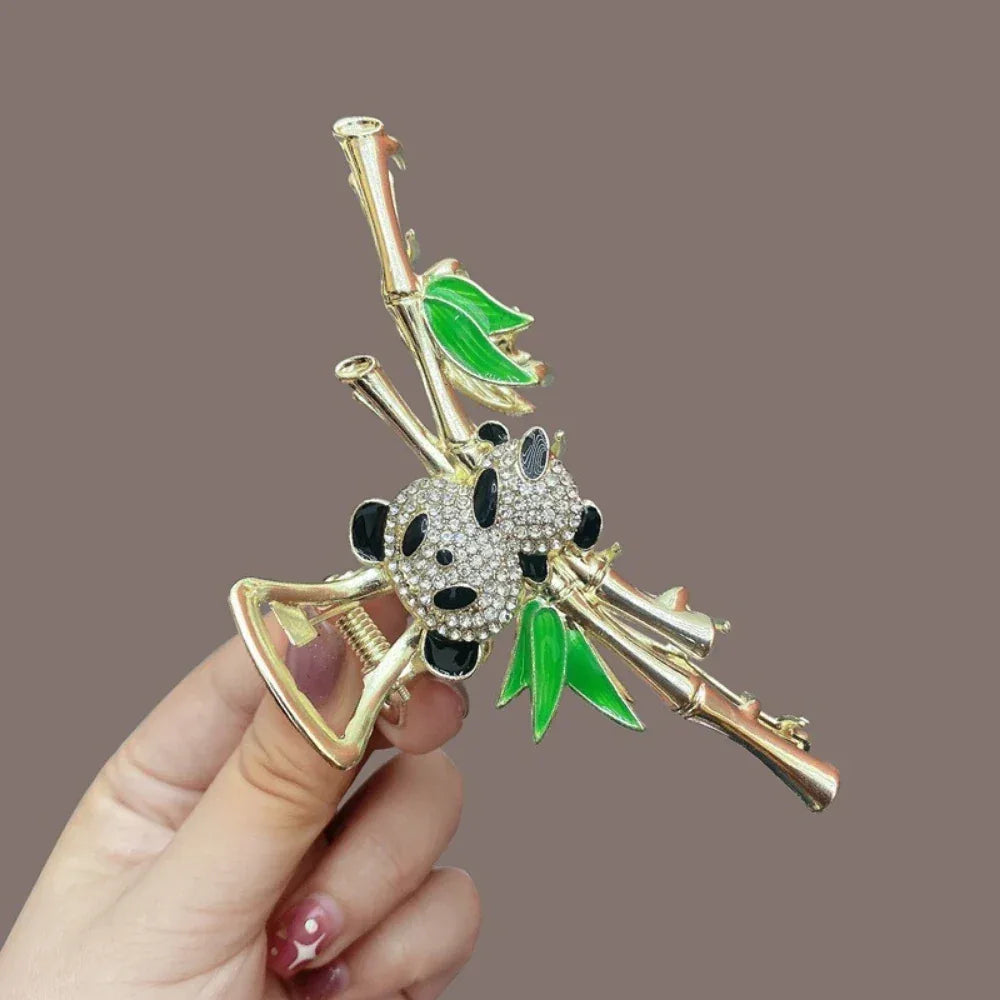 A hand holding a gold claw clip with a panda and bamboo design, showcasing the detail of the rhinestones and enamel.