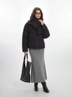 A woman styled in a black puffer jacket, grey skirt, and accessories, standing against a white backdrop.