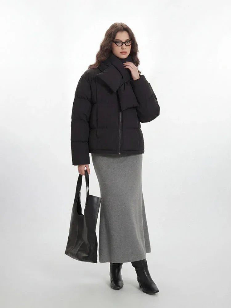 A woman styled in a black puffer jacket, grey skirt, and accessories, standing against a white backdrop.