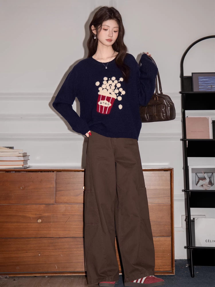 Full body shot of a woman wearing a navy blue knit sweater with a popcorn graphic and Korean text, brown pants, and holding a brown handbag.