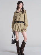 Full body shot of a woman wearing a tan button-down shirt dress, black studded belt, white socks, and brown boots.