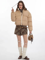Model adjusting hair while wearing a tan cropped puffer jacket, brown plaid pleated mini skirt, white leg warmers, and brown loafers.