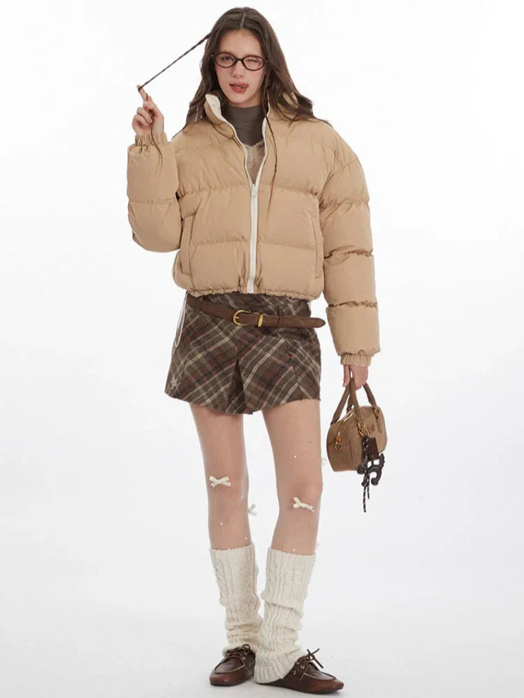 Model adjusting hair while wearing a tan cropped puffer jacket, brown plaid pleated mini skirt, white leg warmers, and brown loafers.