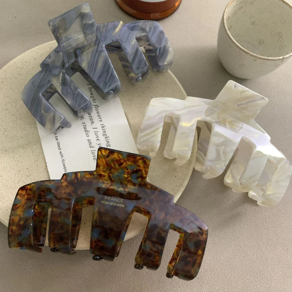 A collection of three claw hair clips in tortoiseshell, grey, and white marbled patterns with a small card and a cup in the background.