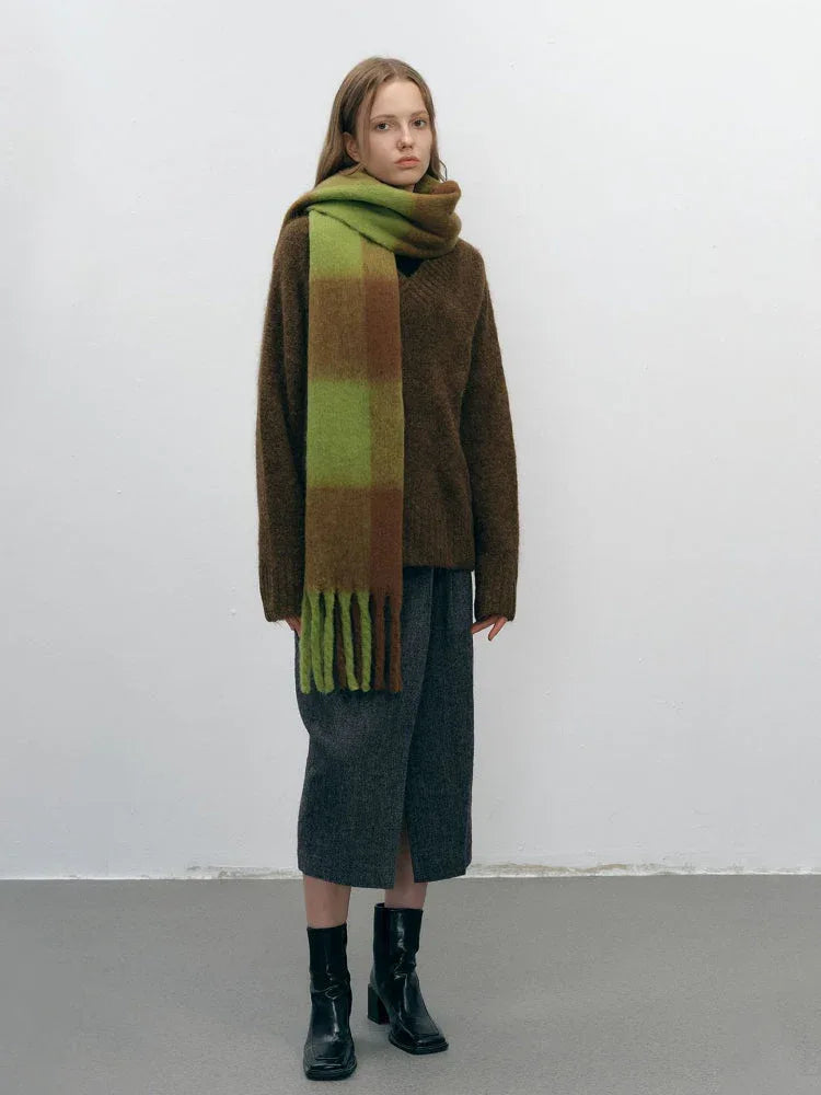 Full body shot of a woman in a brown sweater, grey skirt, black boots, and a green and brown plaid scarf.