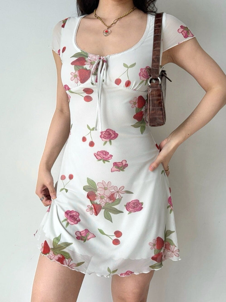 Full body shot of a woman in a white short-sleeve dress with a fruit and floral print, accessorized with a brown handbag and a pendant necklace.