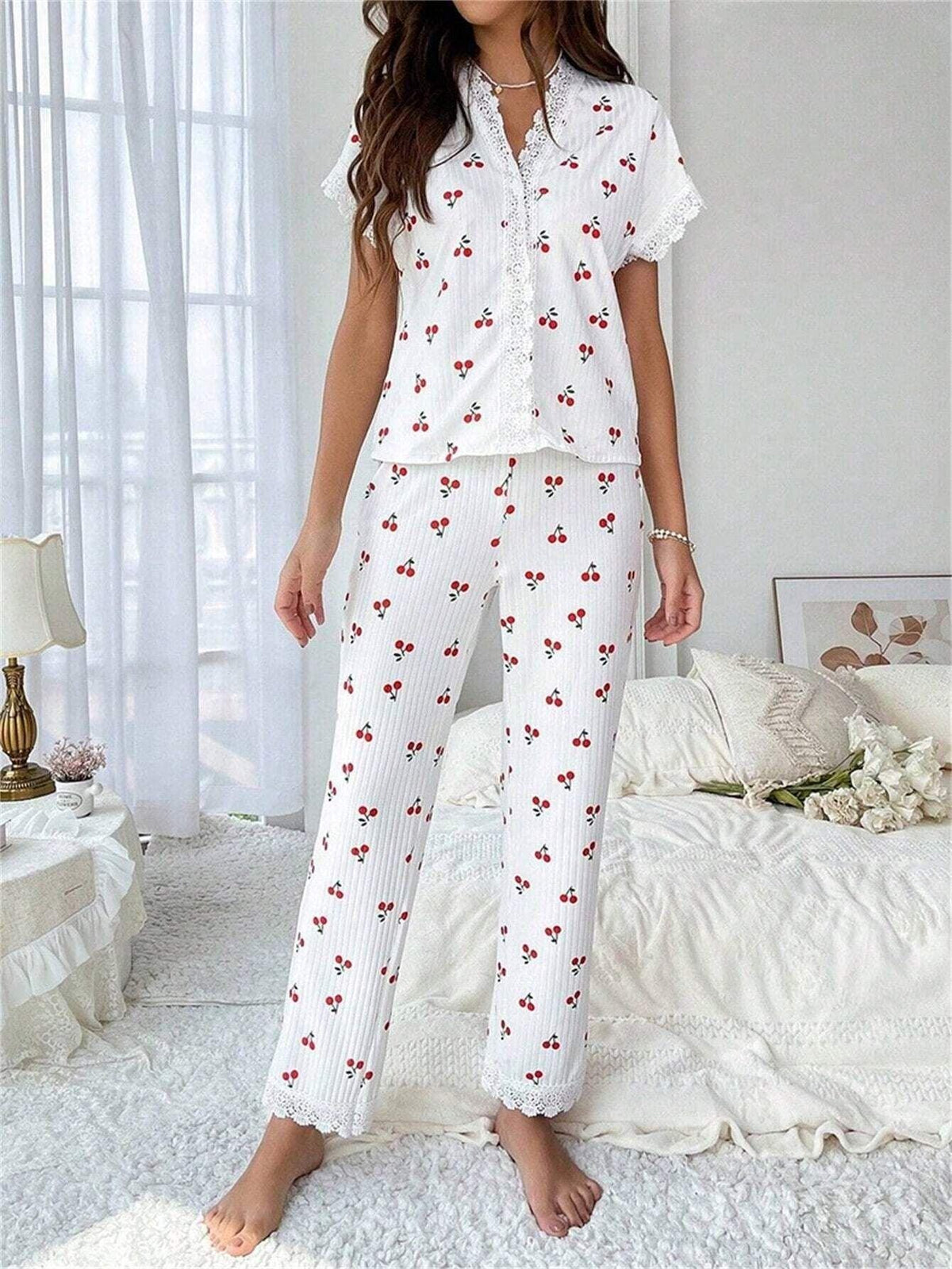 Woman wearing white short-sleeve pajama top with cherry print and white ribbed pants with cherry print, standing in a bedroom. Main image