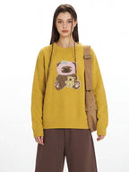 Close-up of a woman wearing a yellow crew neck sweater with an embroidered bear graphic.