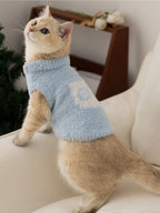 A cream-colored cat wearing a light blue fuzzy sweater is standing on a sofa, looking upwards.