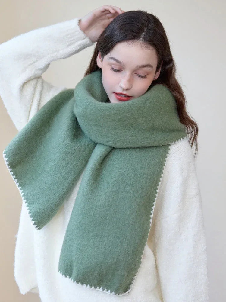 Woman wearing an oversized green scarf with a white fringed edge and a cream-colored sweater, touching her hair.