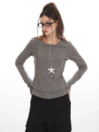 Close-up of a woman wearing a grey long-sleeved shirt, black cargo pants, and a star pendant necklace, holding her glasses.