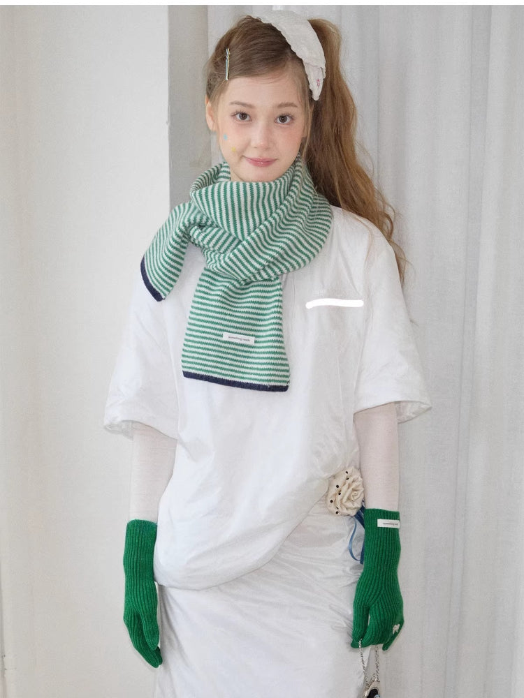 Model wearing a green and white striped knit scarf, white t-shirt, and green knit gloves with bow details, holding charms.