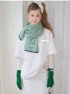 Model wearing a green and white striped knit scarf, white t-shirt, and green knit gloves with bow details, holding charms.