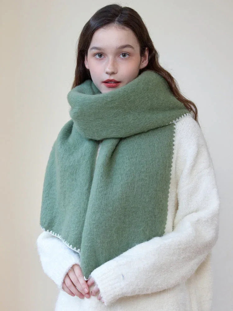 Woman wearing an oversized green scarf with a white fringed edge and a cream-colored sweater, looking forward with scarf draped.