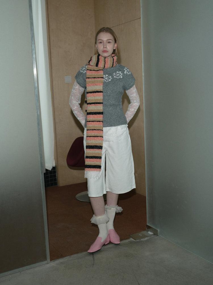 A woman stands wearing a gray sweater with lace sleeves, white shorts, pink shoes, and a striped scarf.