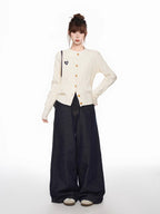 Full body shot of a woman wearing a cream cable knit cardigan and dark wide-leg jeans, standing with one hand on hip