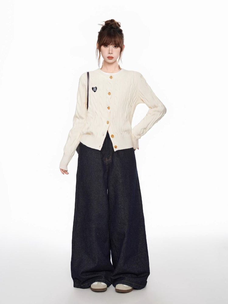 Full body shot of a woman wearing a cream cable knit cardigan and dark wide-leg jeans, standing with one hand on hip