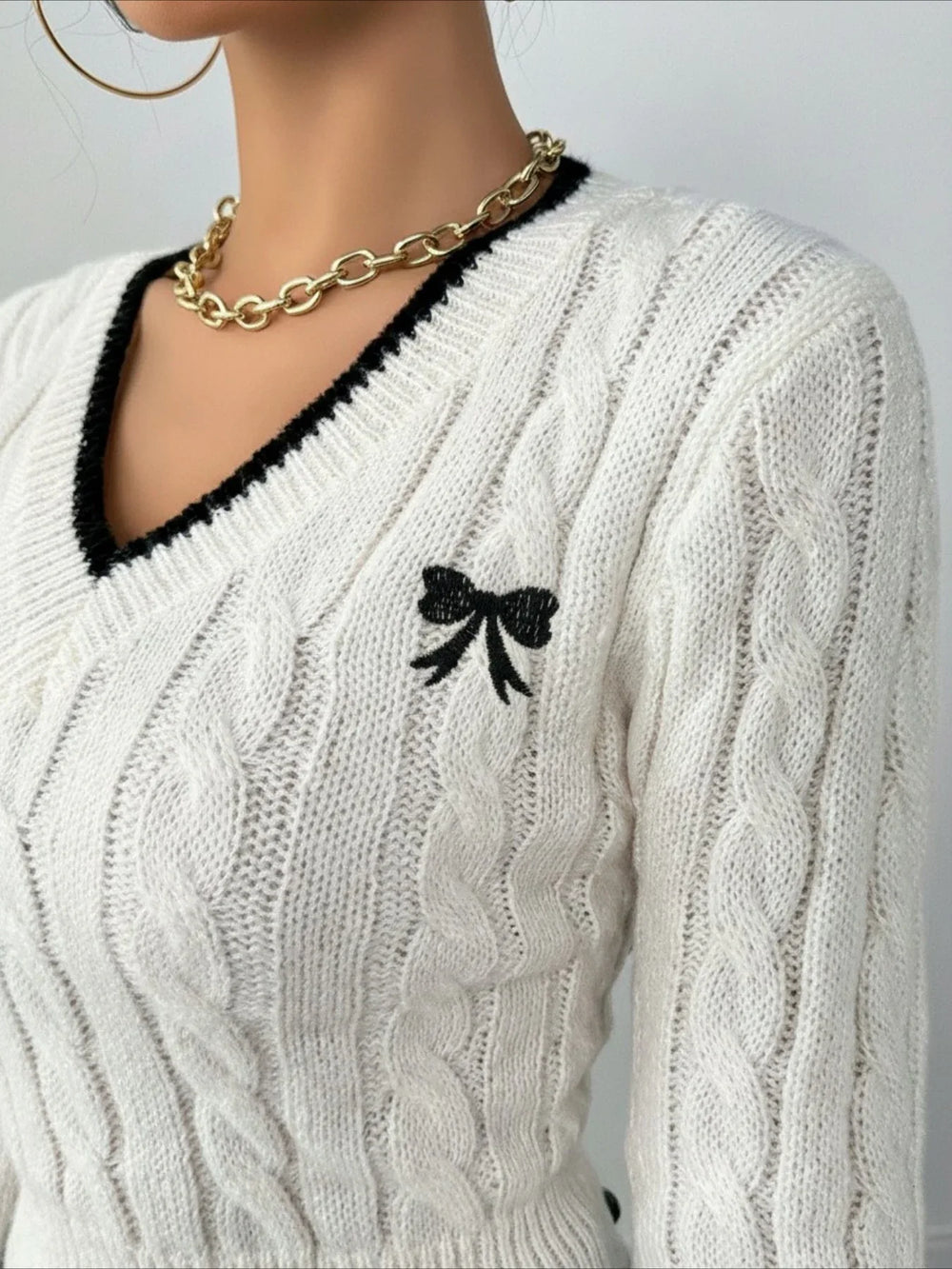 Close-up of the neckline and embroidered bow detail on a white cable knit cropped V-neck sweater with black trim.