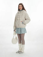 Full body shot of a model wearing a cream fuzzy hooded bomber jacket with toggle closure, a light blue pleated skirt, grey turtleneck, sheer tights, white leg warmers, and a white fuzzy handbag, holding the handbag.