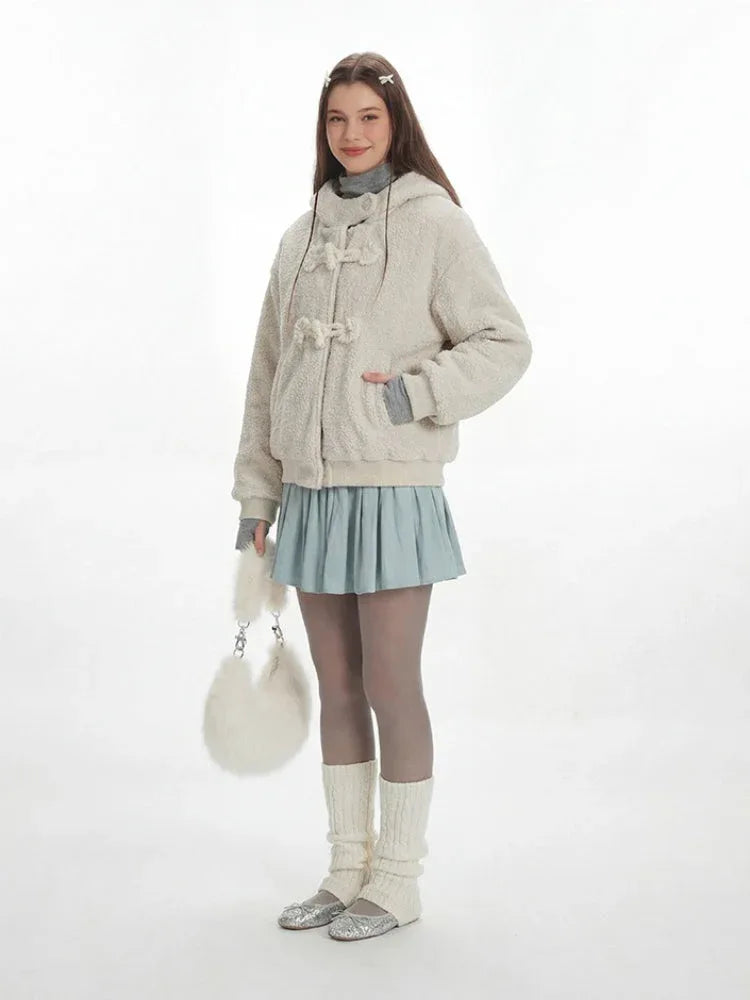 Full body shot of a model wearing a cream fuzzy hooded bomber jacket with toggle closure, a light blue pleated skirt, grey turtleneck, sheer tights, white leg warmers, and a white fuzzy handbag, holding the handbag.
