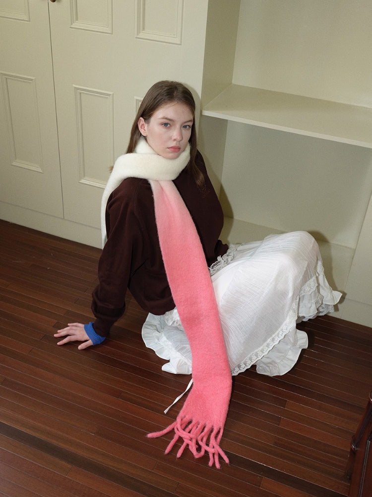 Woman wearing a brown sweatshirt, white ruffled skirt, and a long gradient scarf from cream to pink with fringe detail, sitting on a wooden floor.