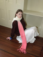 Woman wearing a brown sweatshirt, white ruffled skirt, and a long gradient scarf from cream to pink with fringe detail, sitting on a wooden floor.